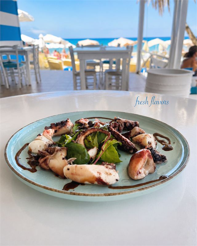 Grilled octopus on baby spinach with a balsamic drizzle, served on a pale blue plate with the beach behind