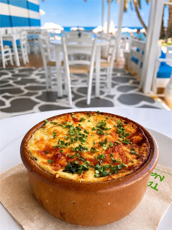 A clay pot of oven-baked cheese, golden on top and sprinkled with chives, with the beach terrace behind
