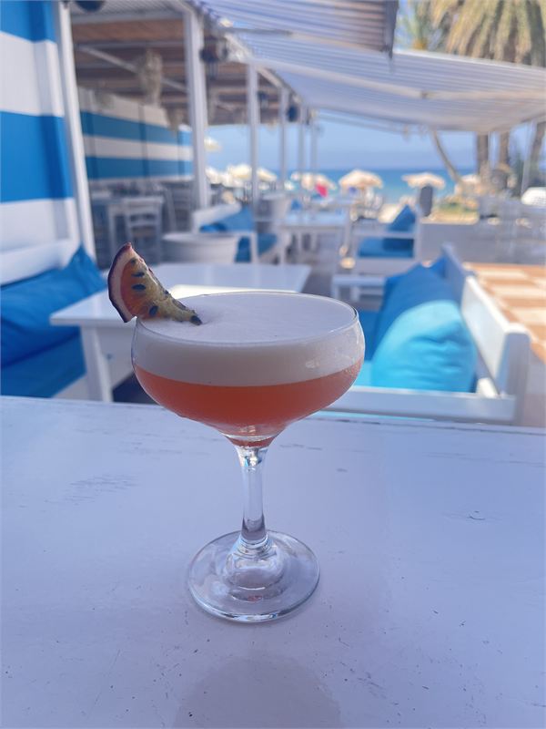 A passion fruit cocktail with a foam top in a coupe glass on a white table, blue cushions and the beach behind