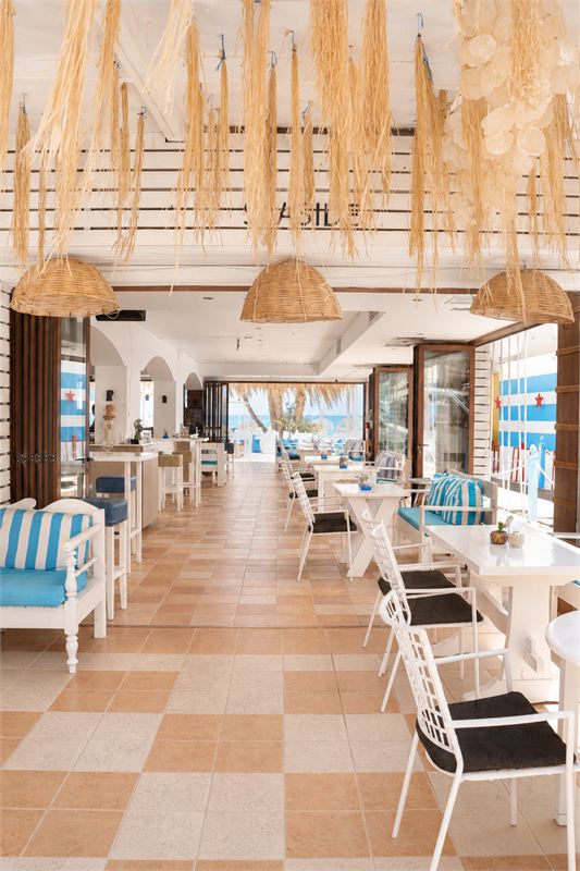 The dining hall with terracotta tiles, white chairs and raffia lamps, opening onto the palms and the sea