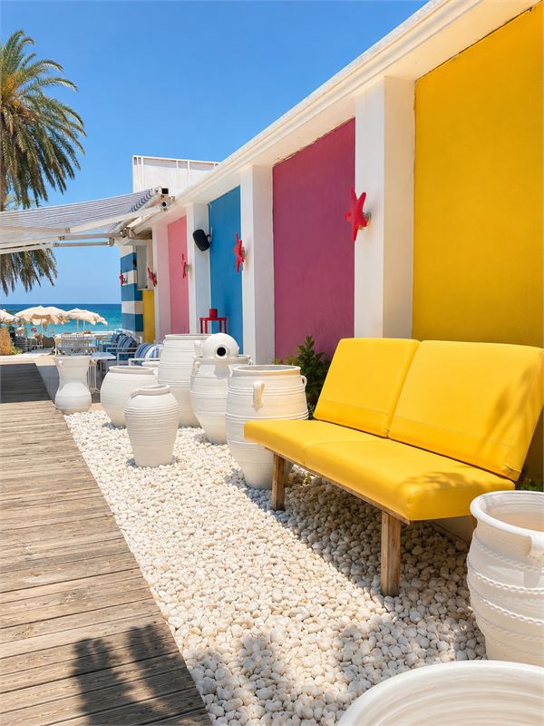 Sunlit terrace with colourful beach cabins, white clay pots on pebbles and a palm tree against a deep blue sky
