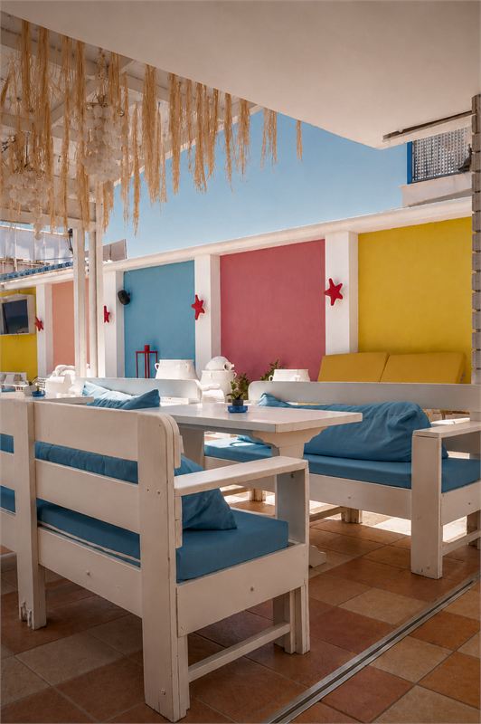 White wooden benches with blue cushions on a terracotta-tiled terrace, colourful cabins and raffia hanging overhead